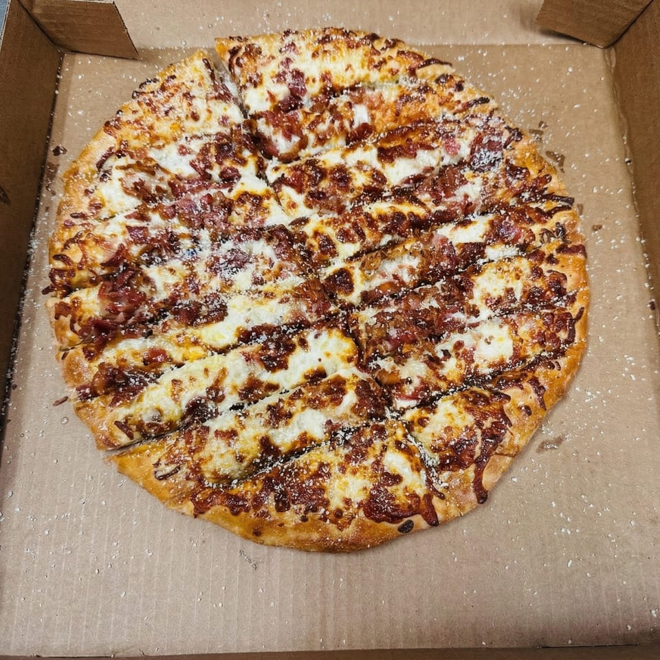 A whole, sliced pizza with white cheese, bacon, and a dark sauce in a cardboard box.