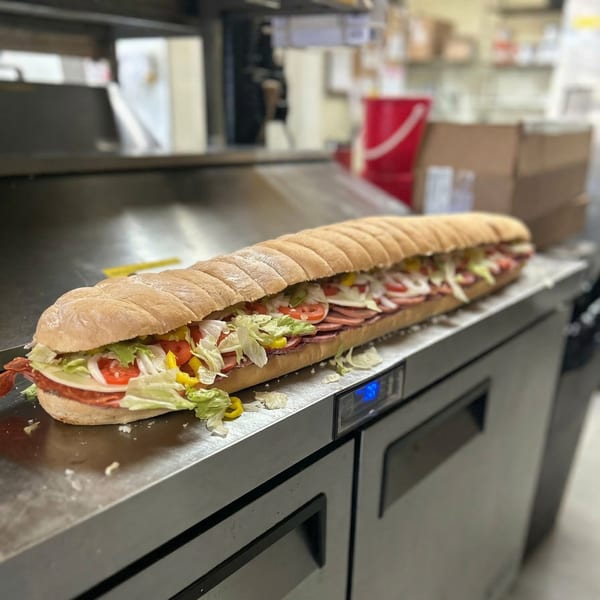 A large, fully loaded sub sandwich with various meats, cheese, and vegetables on a stainless steel counter.