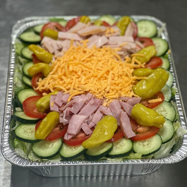 Large catering tray of salad with lettuce, cucumbers, tomatoes, ham, shredded cheese, and peppers.